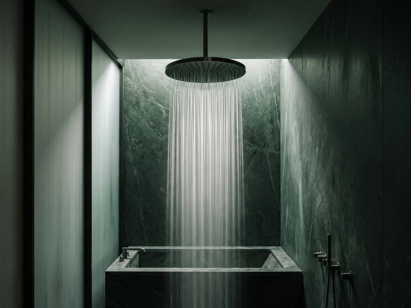 Round rain shower head releasing water strands against dark green stone