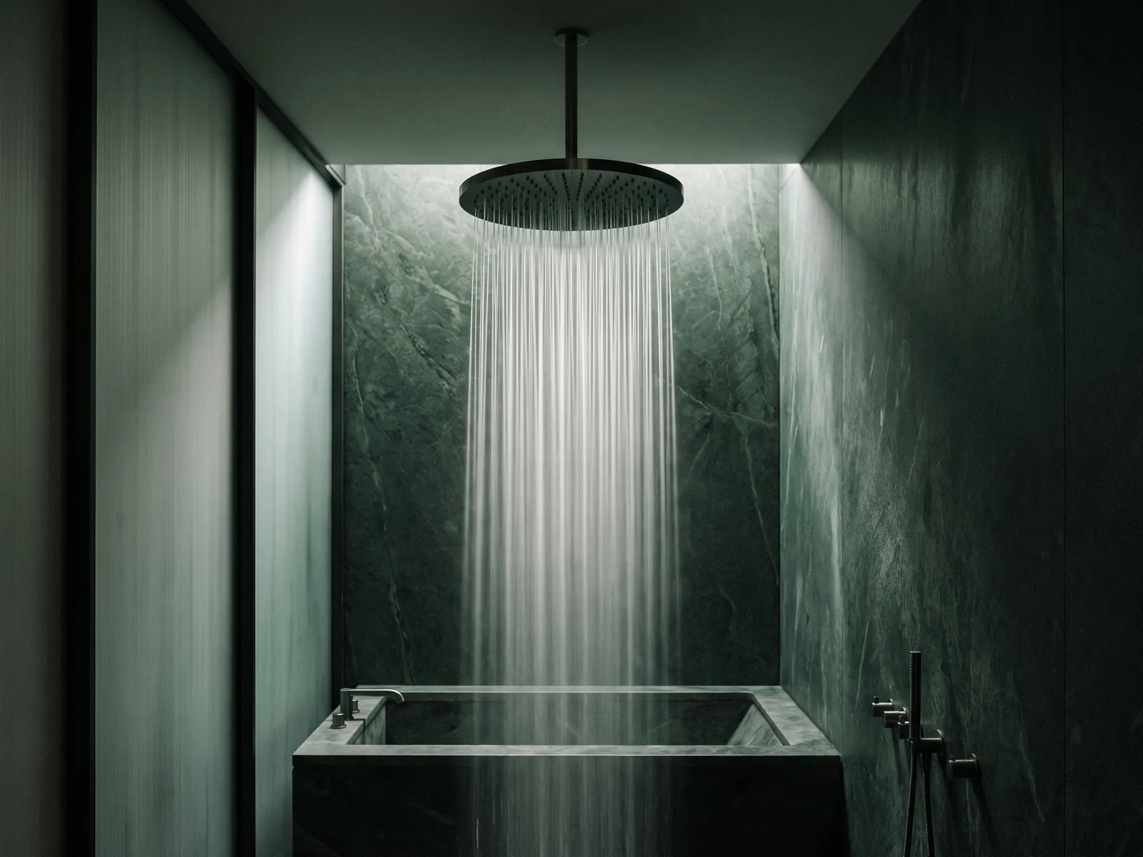 Round rain shower head releasing water strands against dark green stone