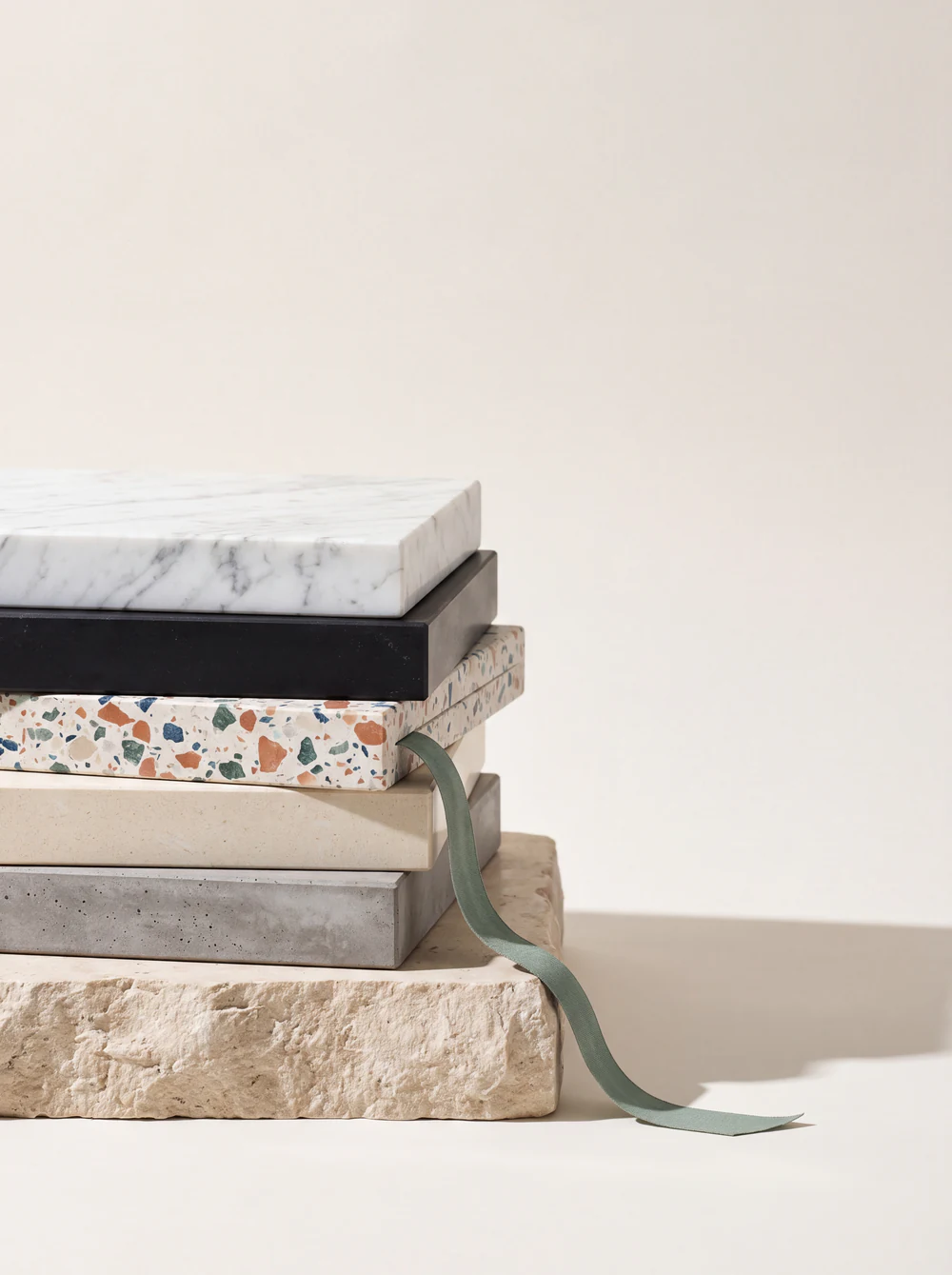 A stack of stone sample slabs - marble, quartz and terrazzo - with a sage linen bookmark, styled like a stack of magazines