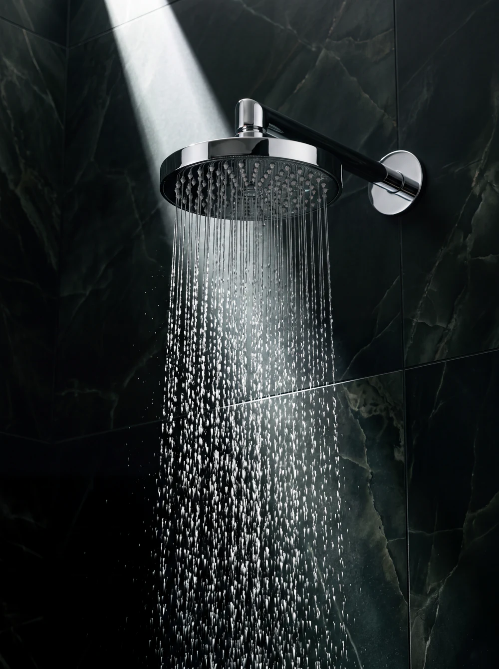 Sculptural chrome rain shower with falling water against dark green tiles
